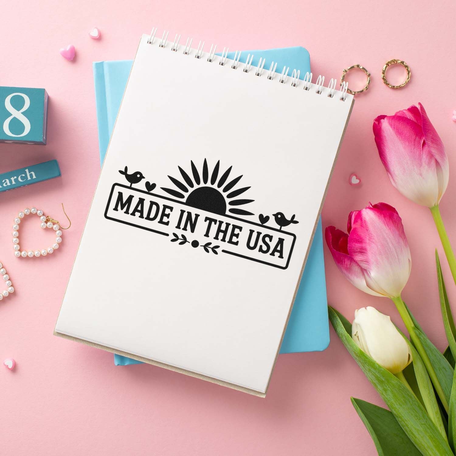 White spiral notebook with black Made in the USA stamp, on a pink background with blue notebooks and tulips.