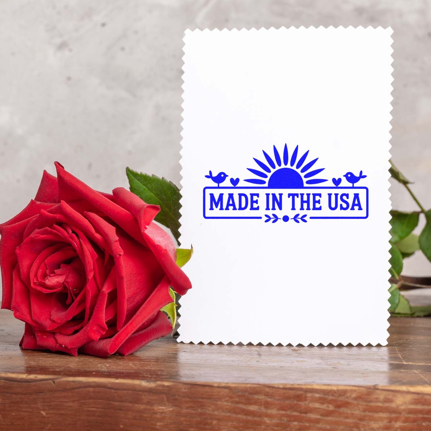 White scalloped card with blue Made in the USA stamp seal beside a red rose on a wooden surface.