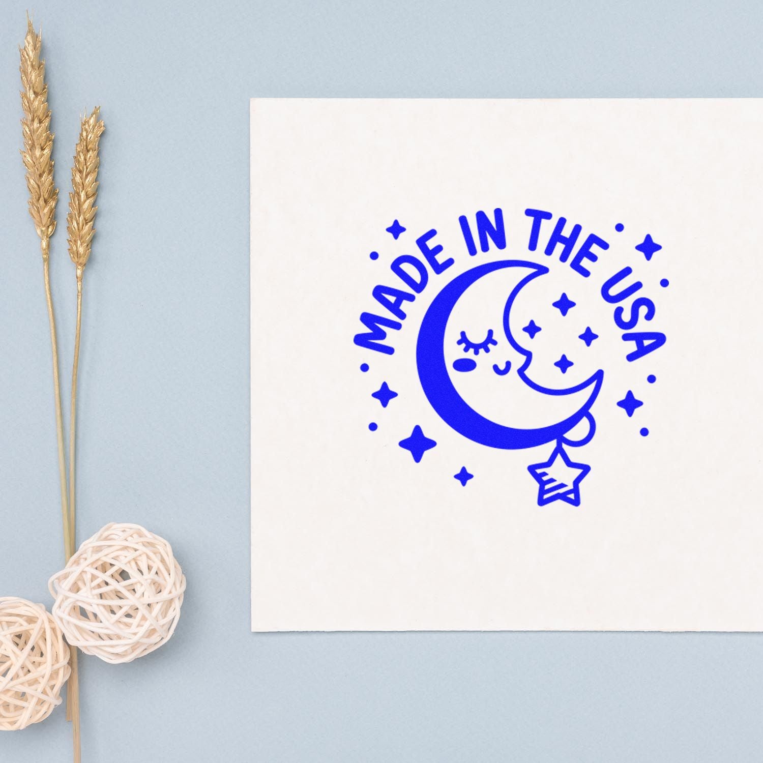 Blue Made in the USA moon and stars stamp seal on white paper, with gold wheat stems and woven balls on a pale blue background.