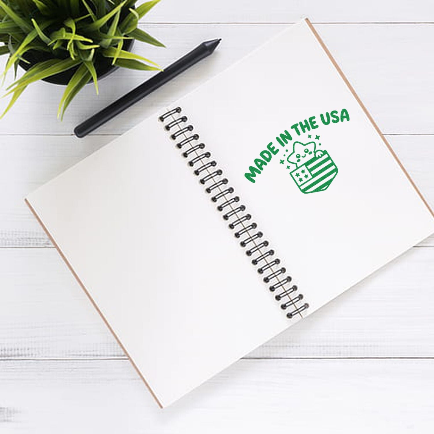 White spiral notebook with green Made in the USA cat stamp and seal, plus black pen and plant.