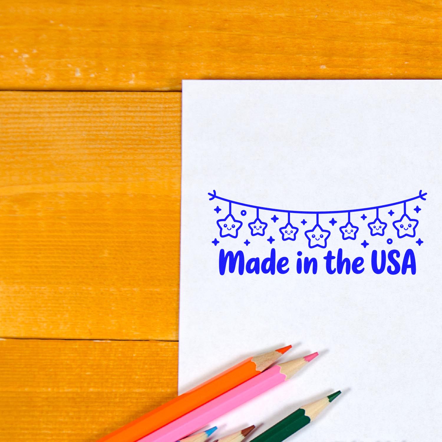 Blue Made in the USA stamp/seal with smiling star garland on white paper, plus colored pencils on a wooden desk.