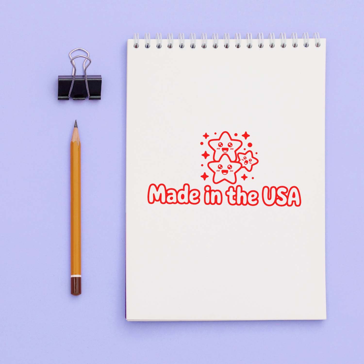 White spiral notebook with red Made in the USA stamp seal, beside a pencil and black binder clip on a lavender background.