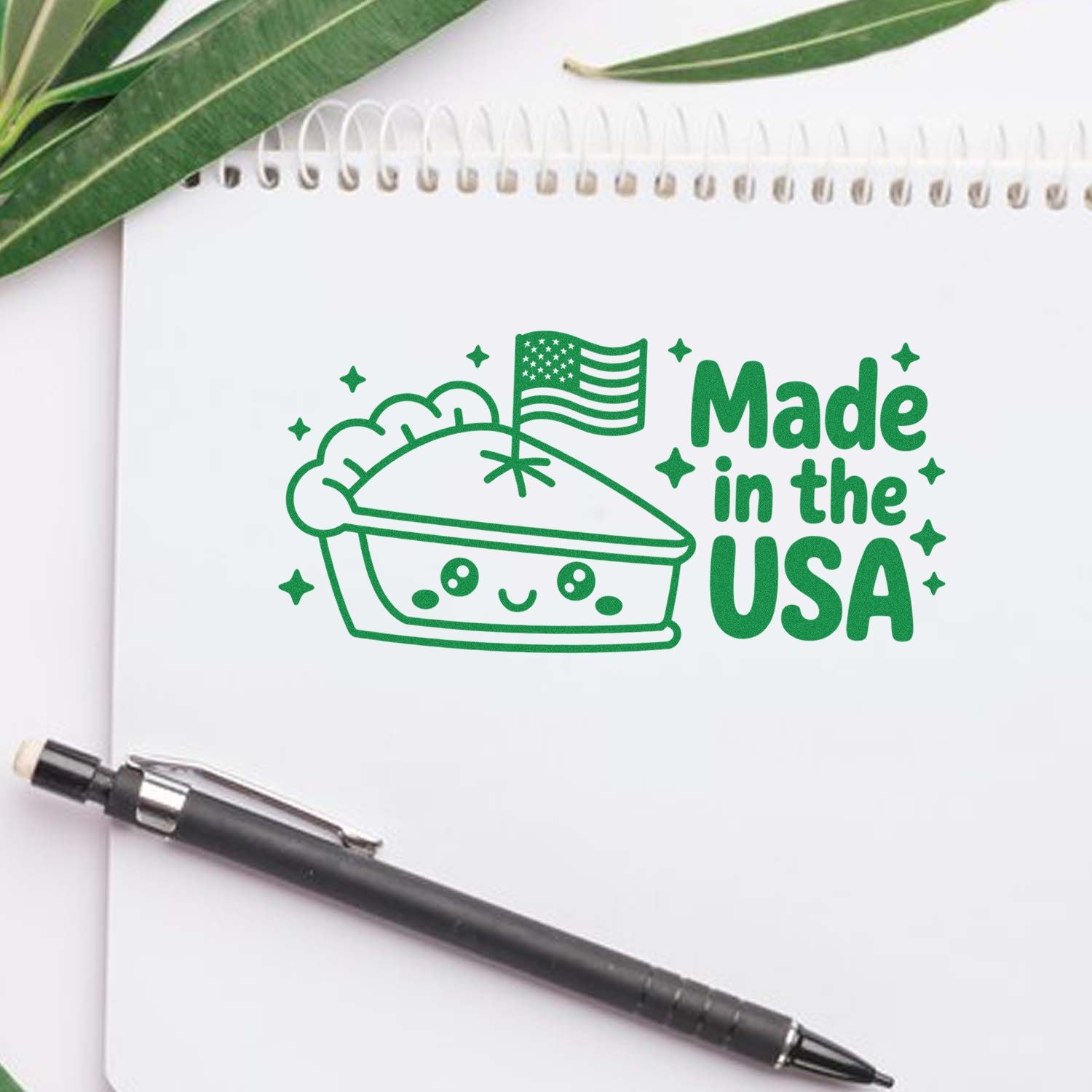 Green Made in the USA stamp seal with patriotic pie icon on a white notebook, beside a black pen and green leaves.