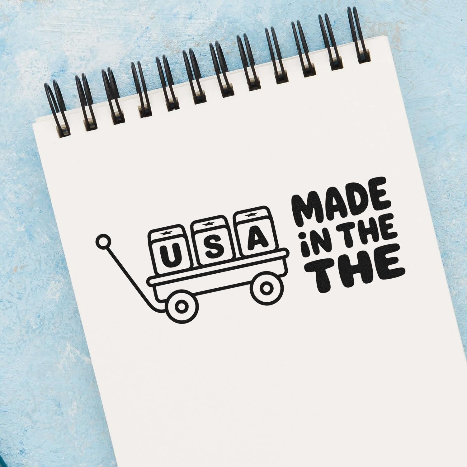 Black Made in the USA stamp seal on a white spiral notebook, with wagon and blue textured background.