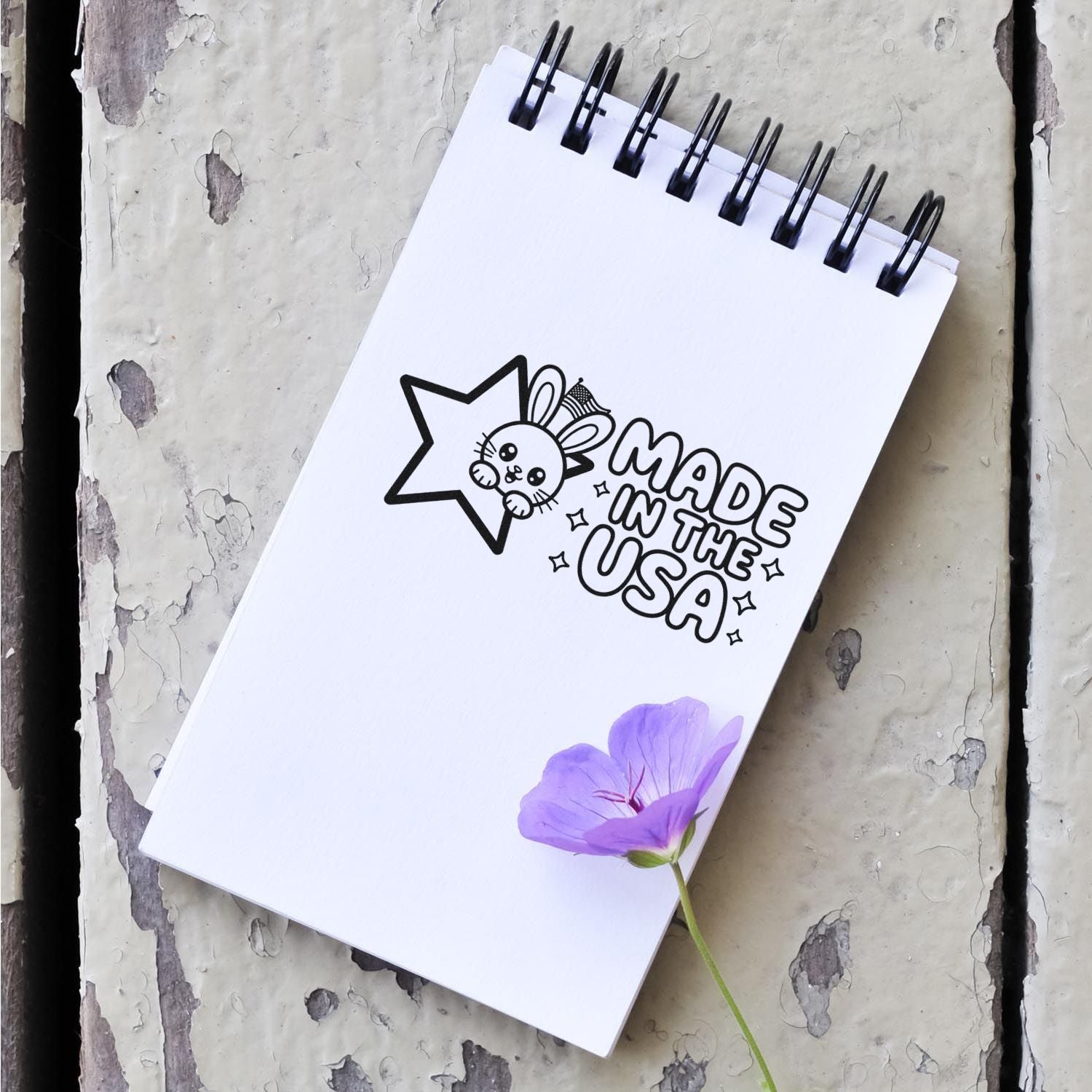 White spiral notebook with black Made in the USA stamp-style seal, bunny star logo, and purple flower on distressed gray wood.