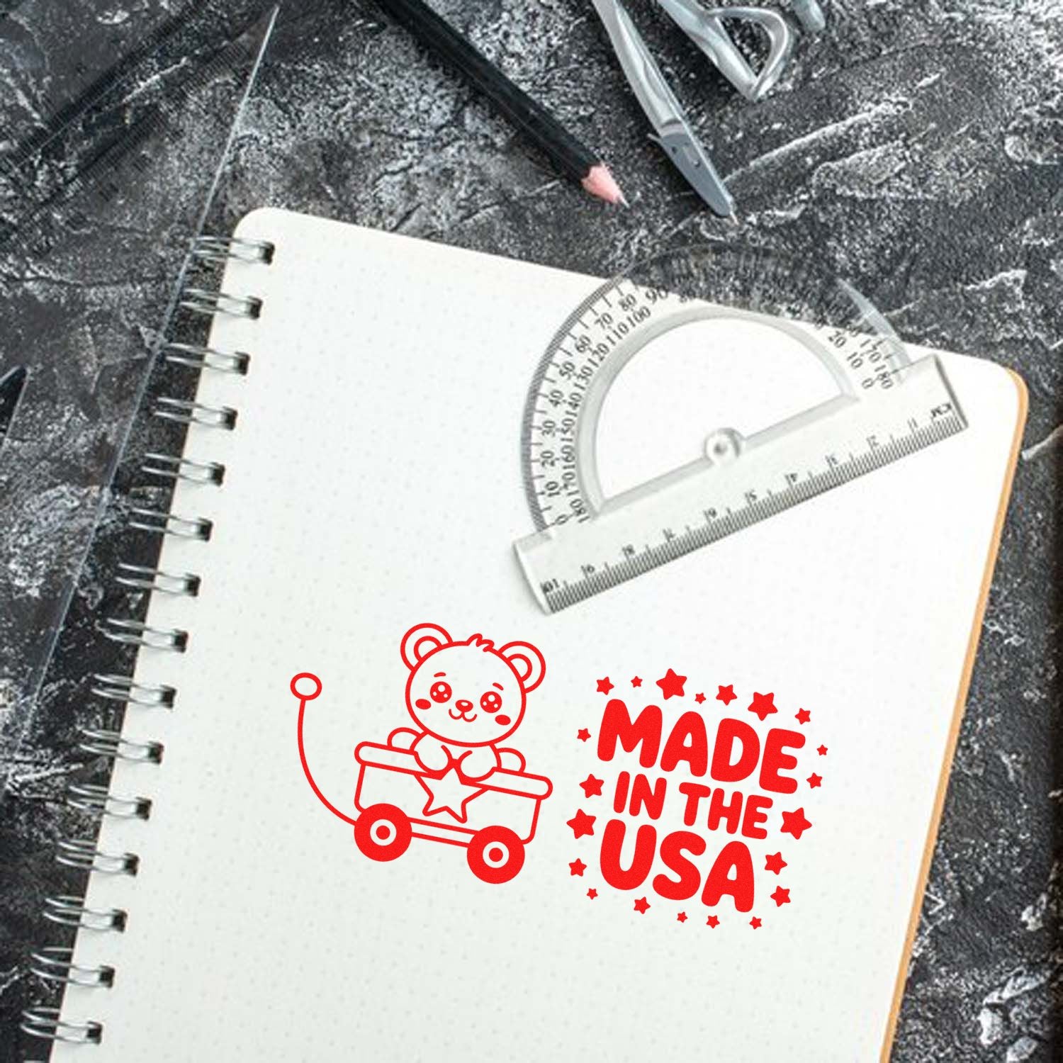 Red custom stamp and seal designs on a spiral notebook: teddy bear cart and Made in the USA in bold red, with drafting tools.