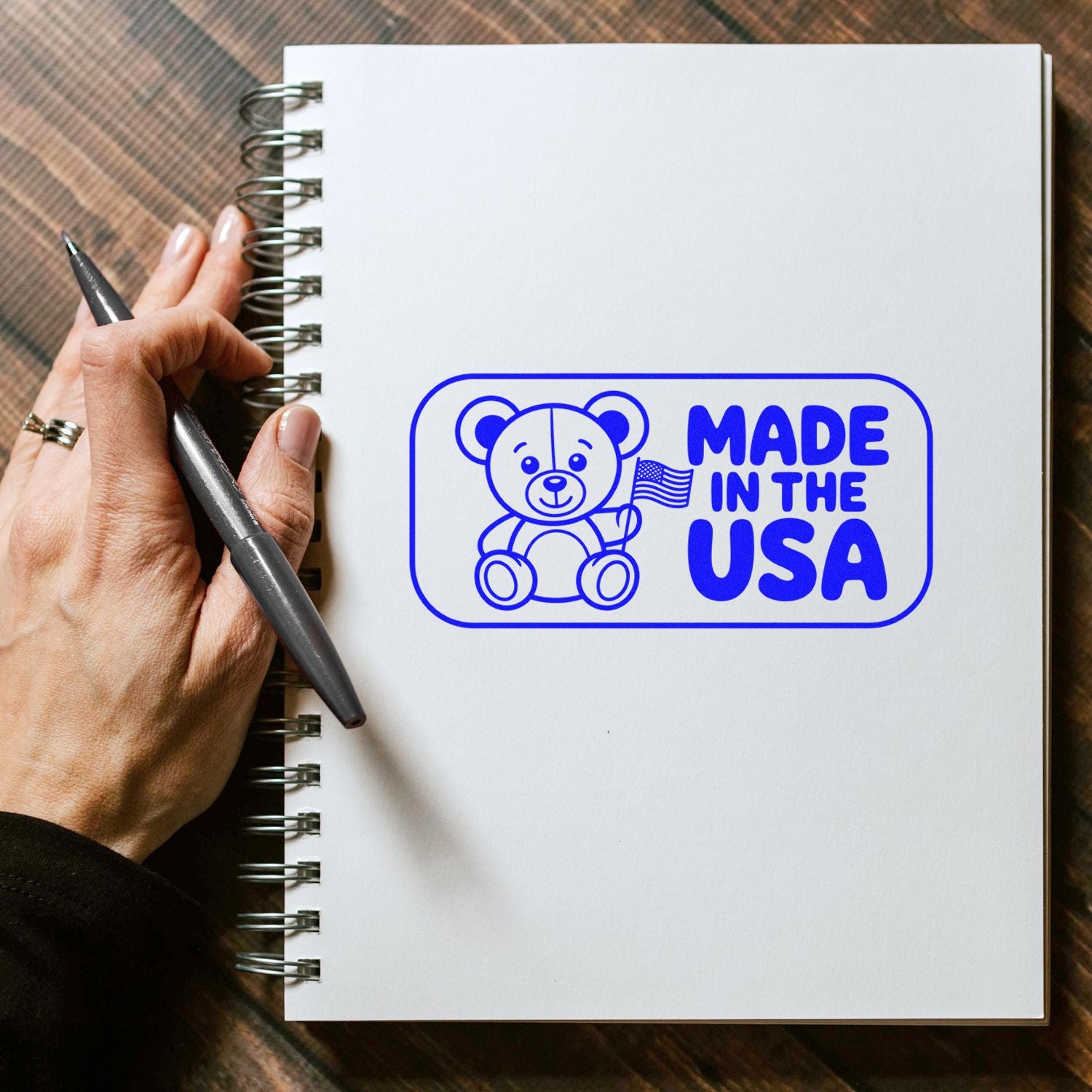 Blue Made in the USA teddy bear stamp seal on white spiral notebook, held by hand with pen.