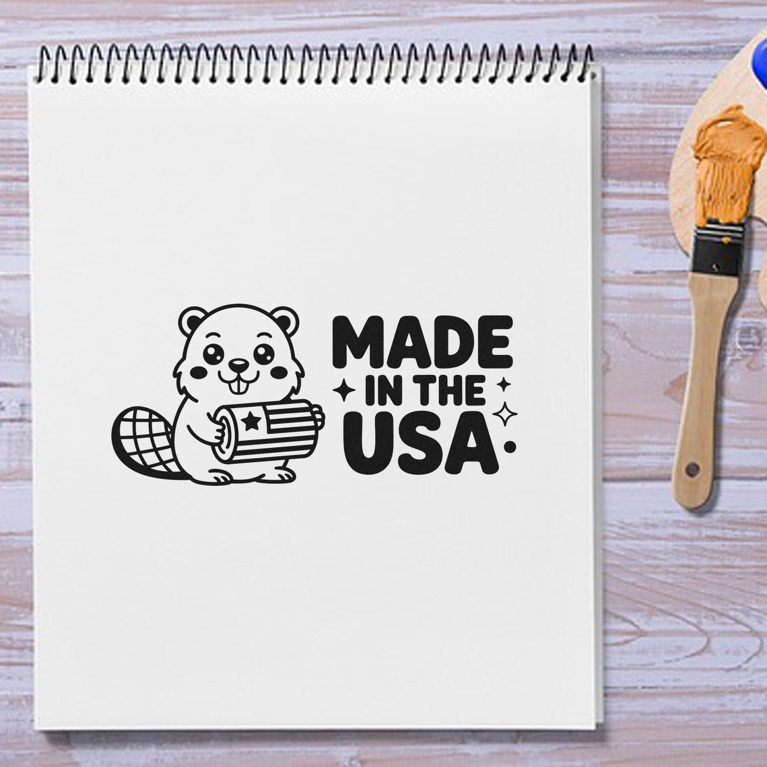 Black Made in the USA beaver stamp design on a white sketchbook, with a wooden stamp tool and orange seal brush.