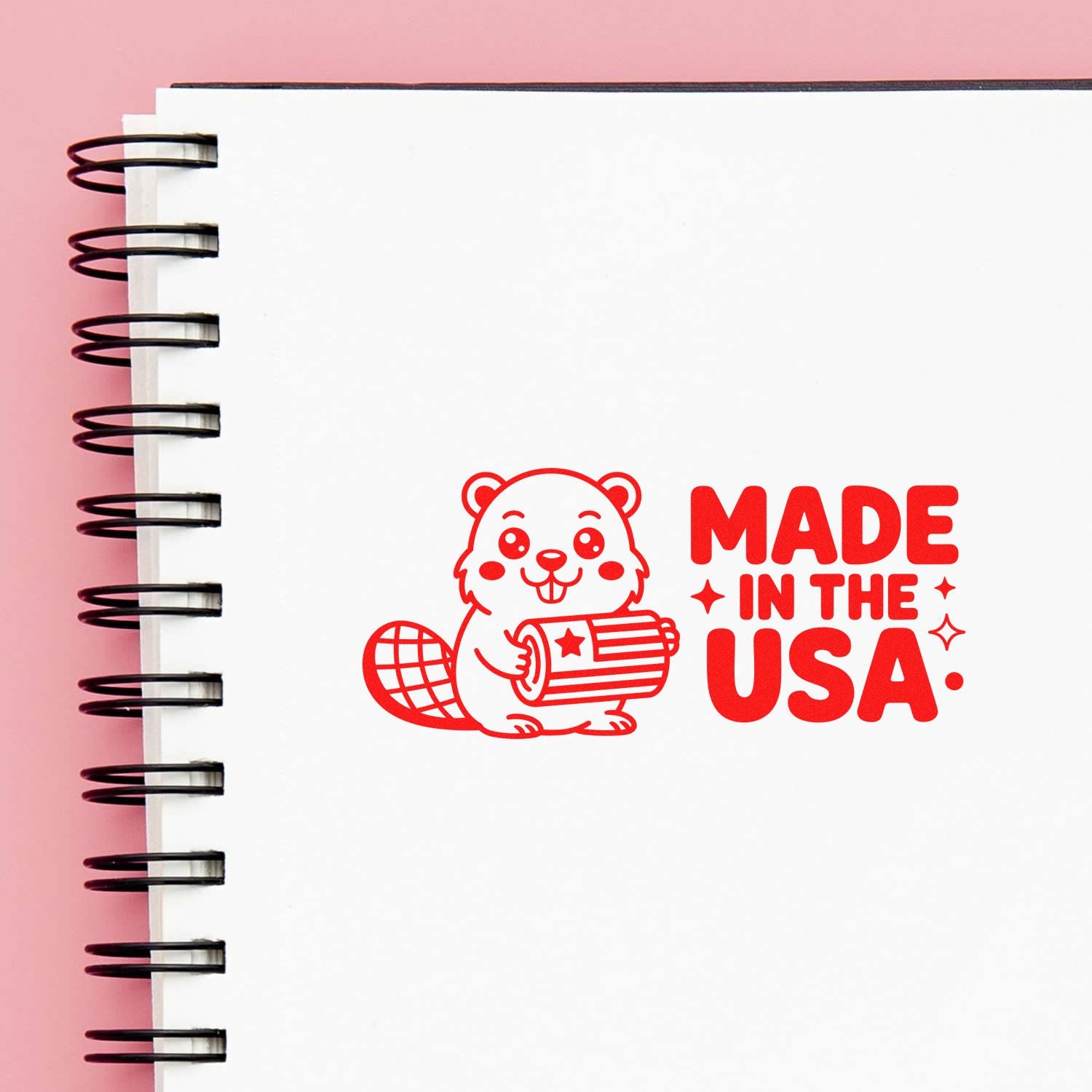 Red stamp-style seal with beaver holding a barrel and Made in the USA on a white spiral notebook over pink.