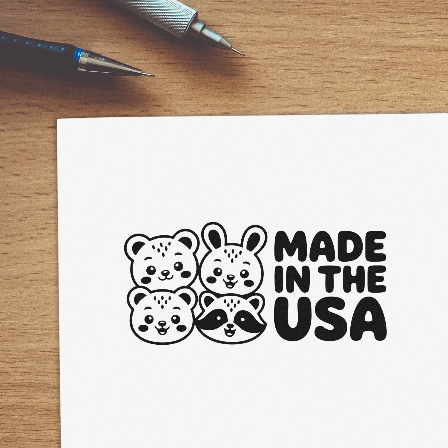 Black Made in the USA stamp with cute bear, bunny, and raccoon faces on white paper, beside pens on a wood desk.