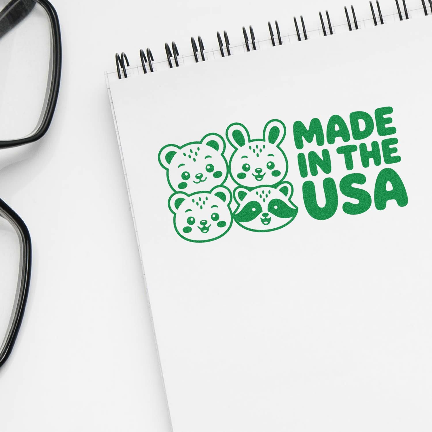 Green Made in the USA stamp and seal design with cute animal faces on a white spiral notebook, beside black glasses.