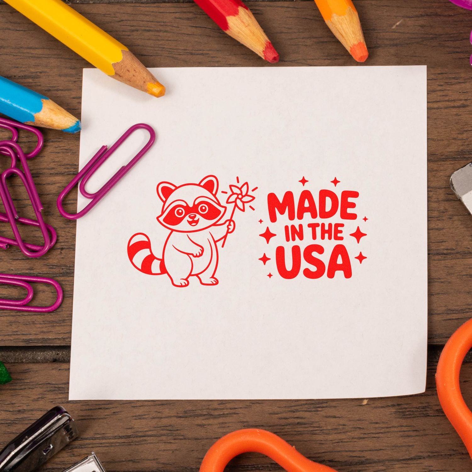 Red Made in the USA raccoon stamp and seal on white paper, surrounded by colorful pencils and magenta paper clips.