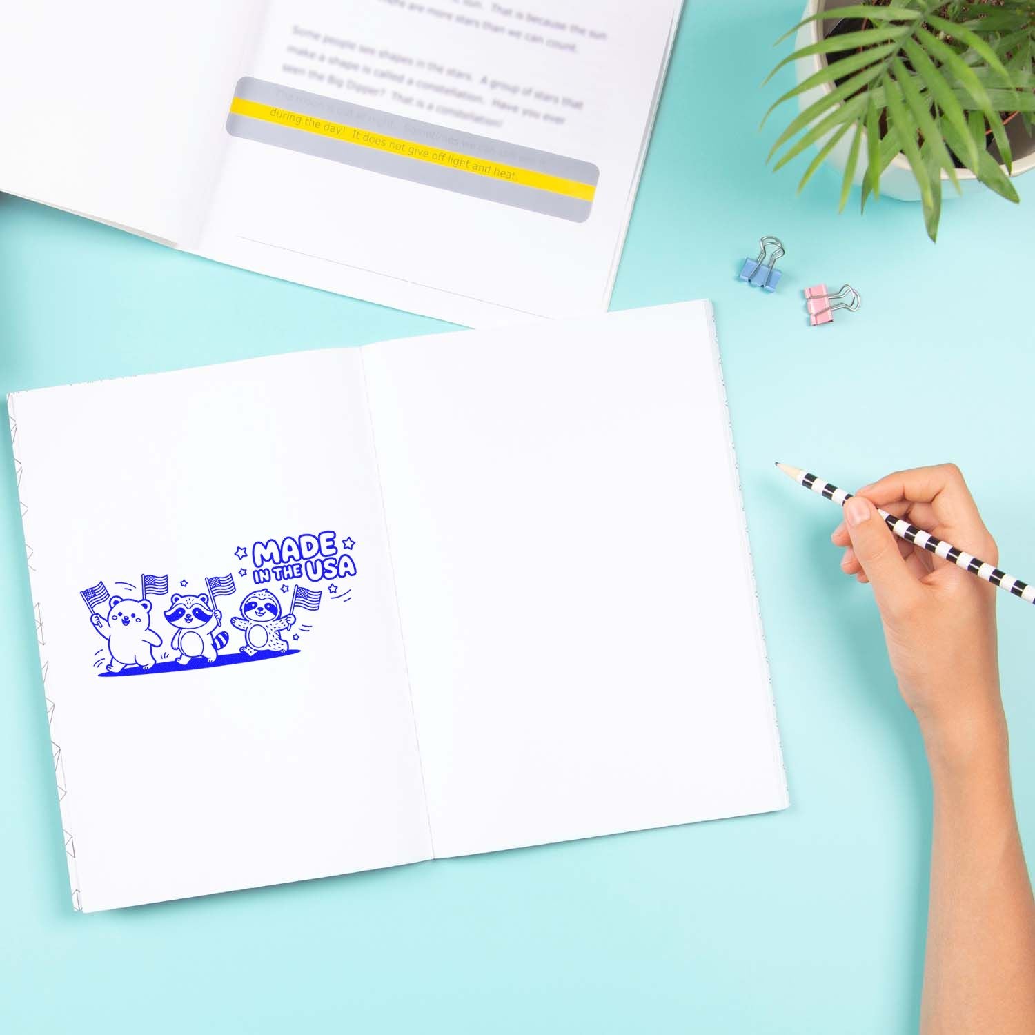 Blue stamp and yellow seal impressions on open notebook, with hand holding a striped pencil on an aqua desk.