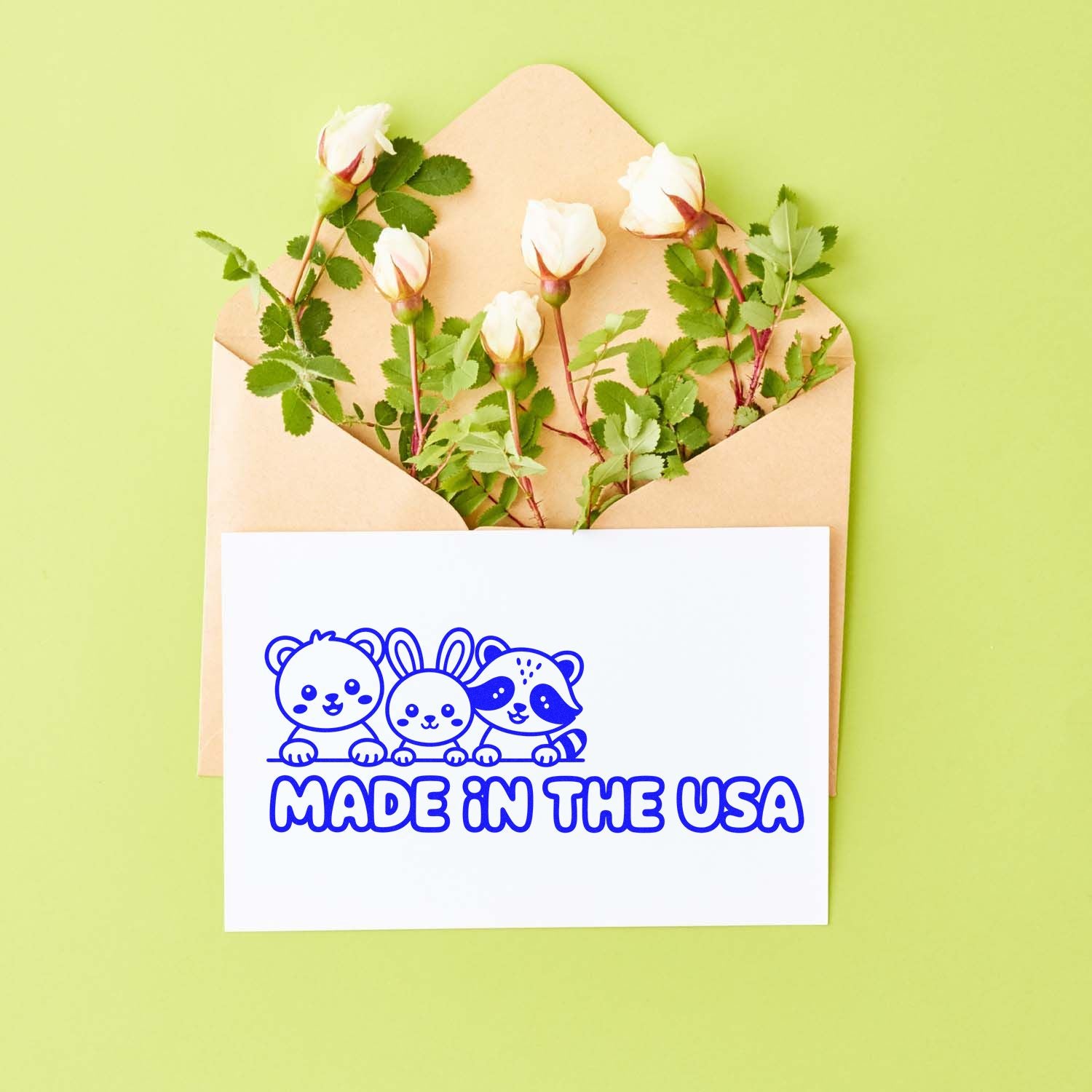 Blue Made in the USA stamp seal with cartoon animals on white card, tucked in a beige envelope with white roses on lime green background.