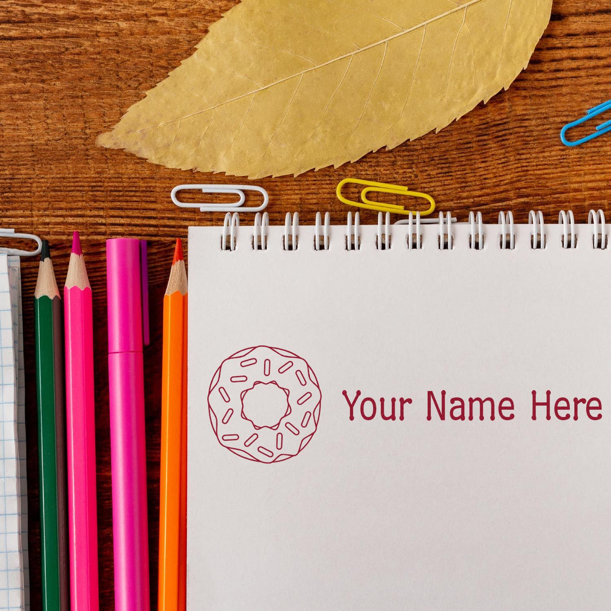 Custom stamp and seal with donut logo and Your Name Here in burgundy on white notebook, beside neon pencils and clips.