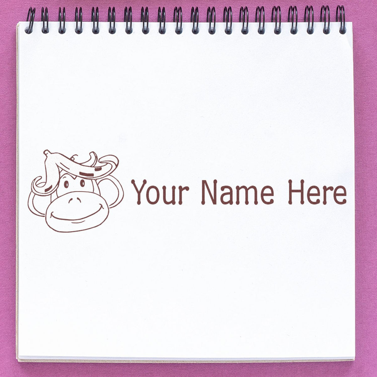 White spiral notebook with a brown custom name stamp, monkey logo, and Your Name Here on a pink background.