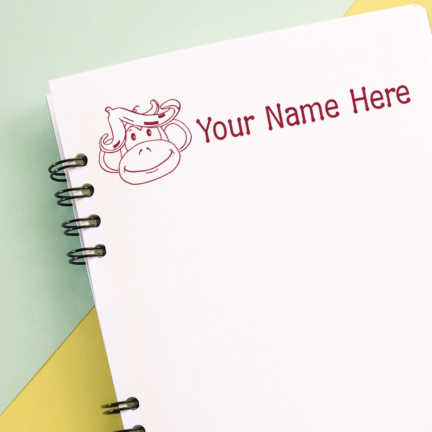 Burgundy custom cow stamp and seal with Your Name Here on a white spiral notebook page, over pastel green and yellow paper.