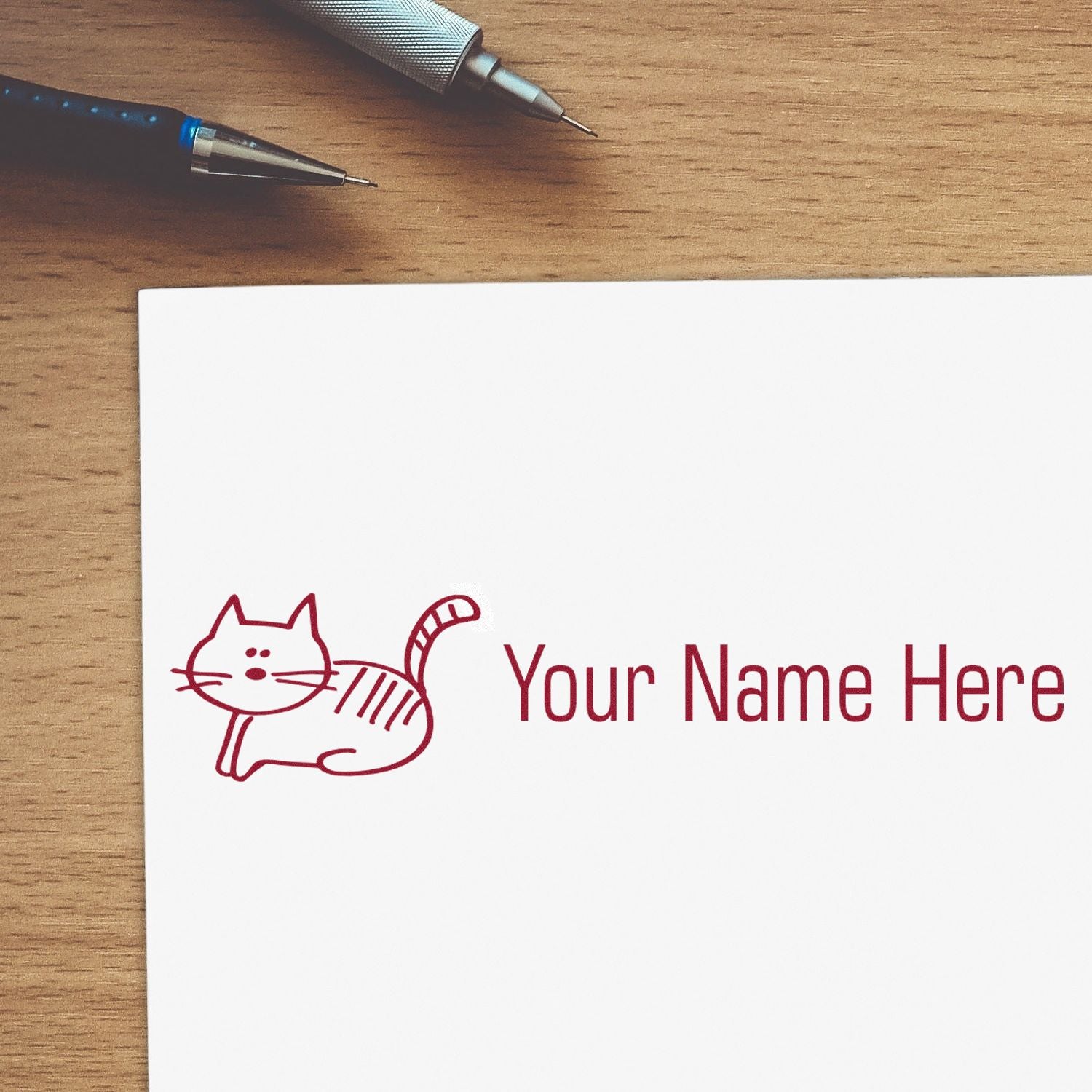 Burgundy custom stamp and seal with cat logo and Your Name Here on white paper, with pens on a wood desk.