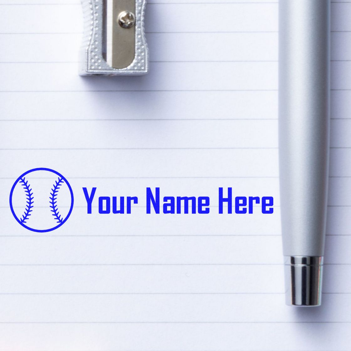 Blue baseball custom stamp and seal with Your Name Here on lined paper beside a silver pen and sharpener.
