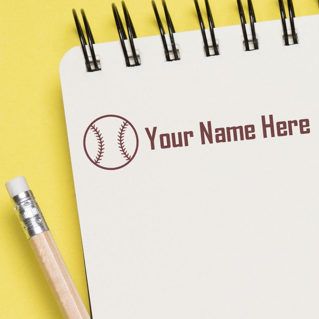 Custom red-brown baseball stamp and seal on a white spiral notebook with yellow background and pencil.