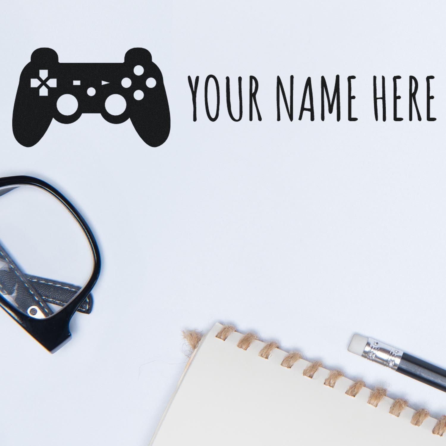 Black custom stamp seal with game controller icon and Your Name Here on white background, plus notebook and pen accents.