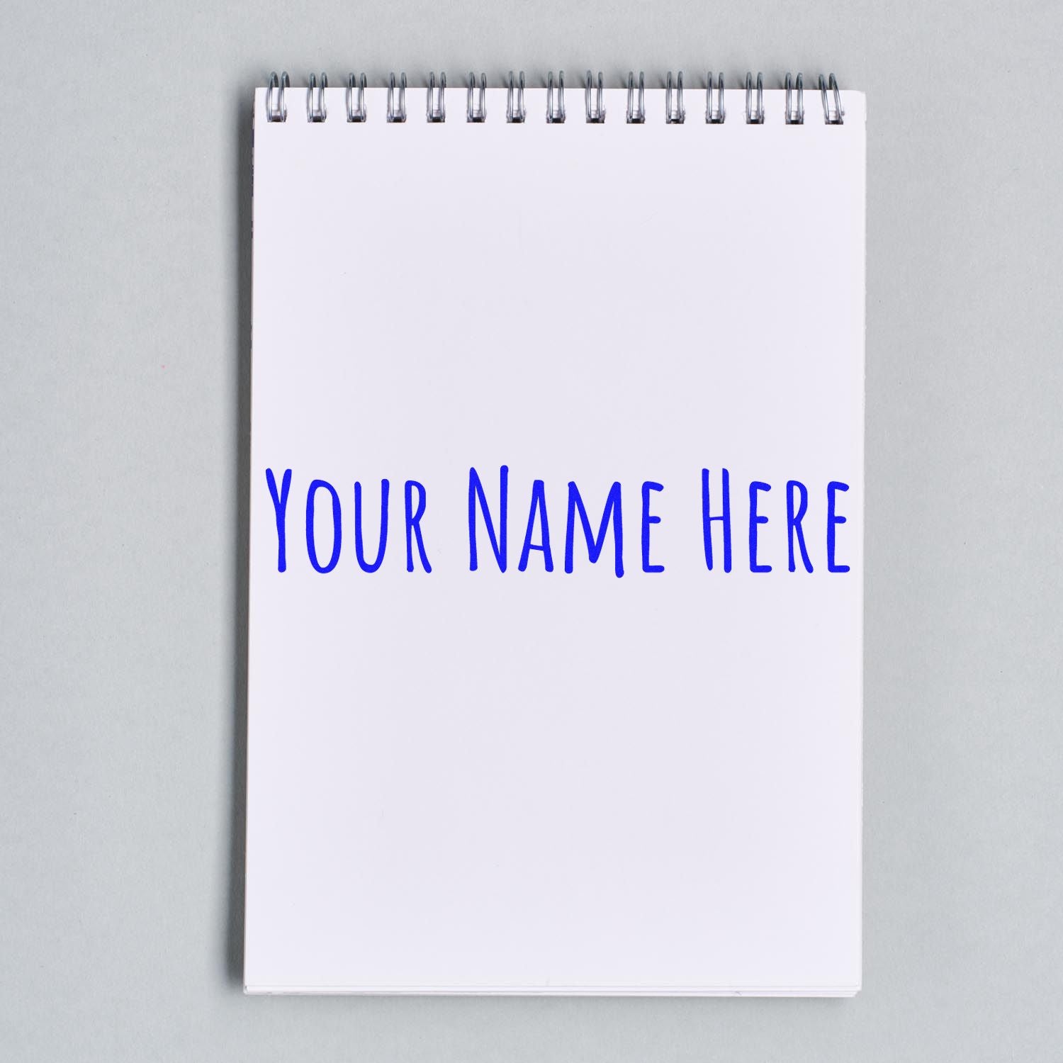 Custom stamp and seal mockup: blue YOUR NAME HERE on a white spiral notebook with a light gray background.