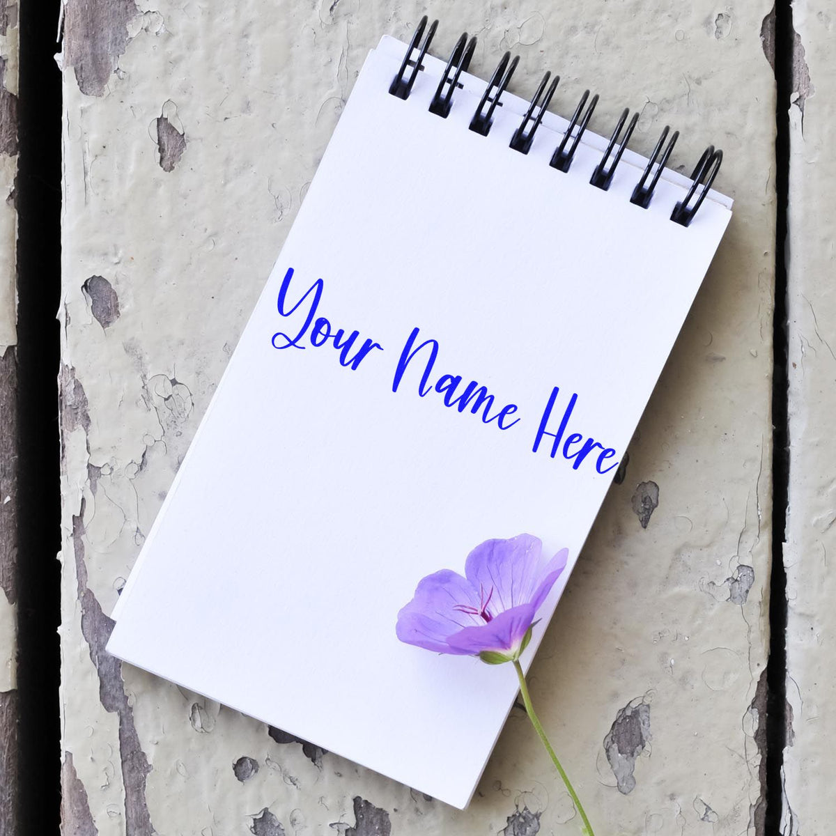 Custom stamp and seal notepad with blue name text, black spiral binding, and purple flower on weathered gray wood.
