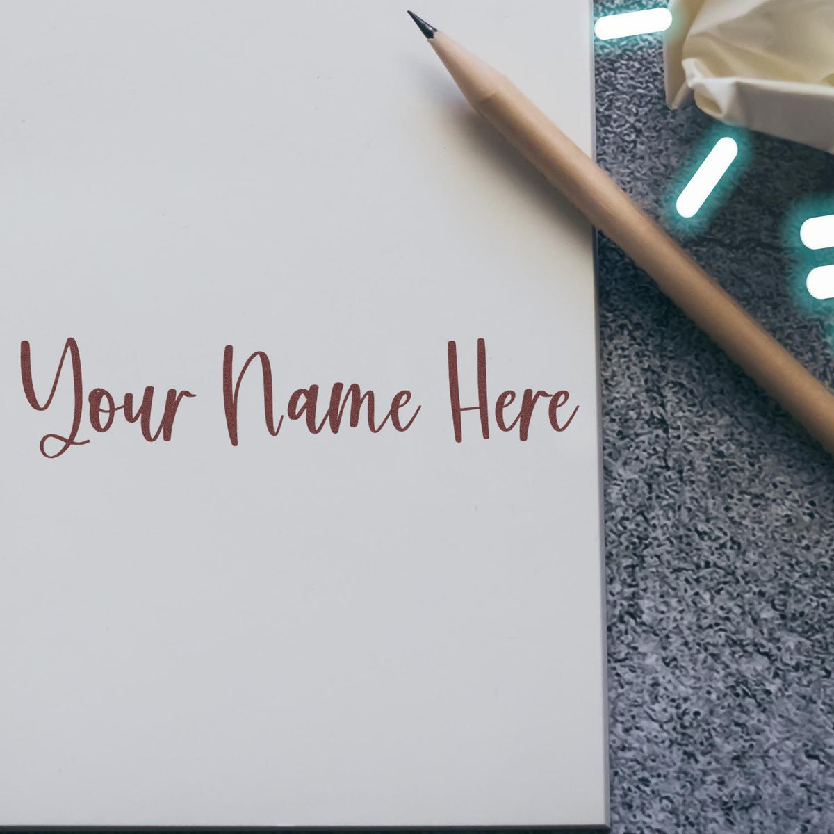 White custom stamp and seal sample with maroon ÂYour Name Here script, beside a wooden pencil on gray textured surface.