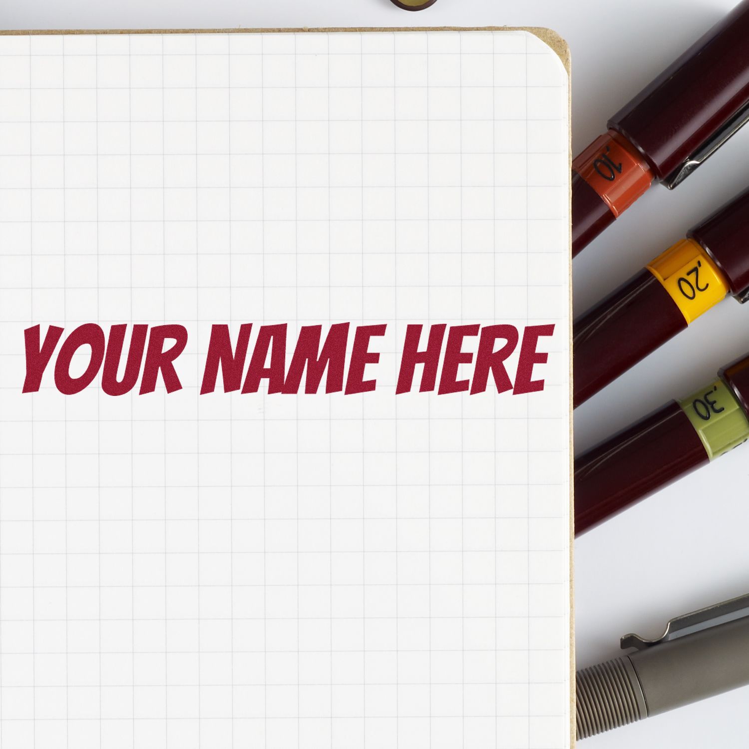 Grid notebook with red YOUR NAME HERE and burgundy stamp and seal pens with orange, yellow, green, and gray bands.