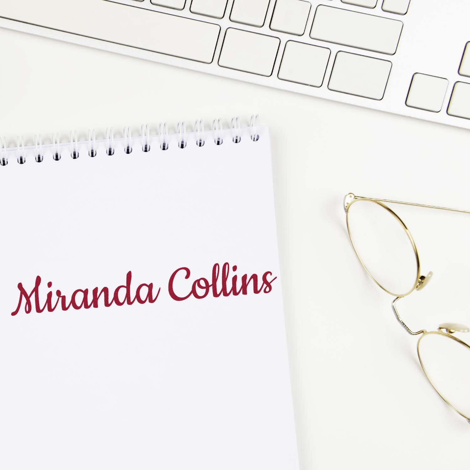 Maroon Miranda Collins custom stamp and seal on white spiral notebook beside gold eyeglasses and keyboard.