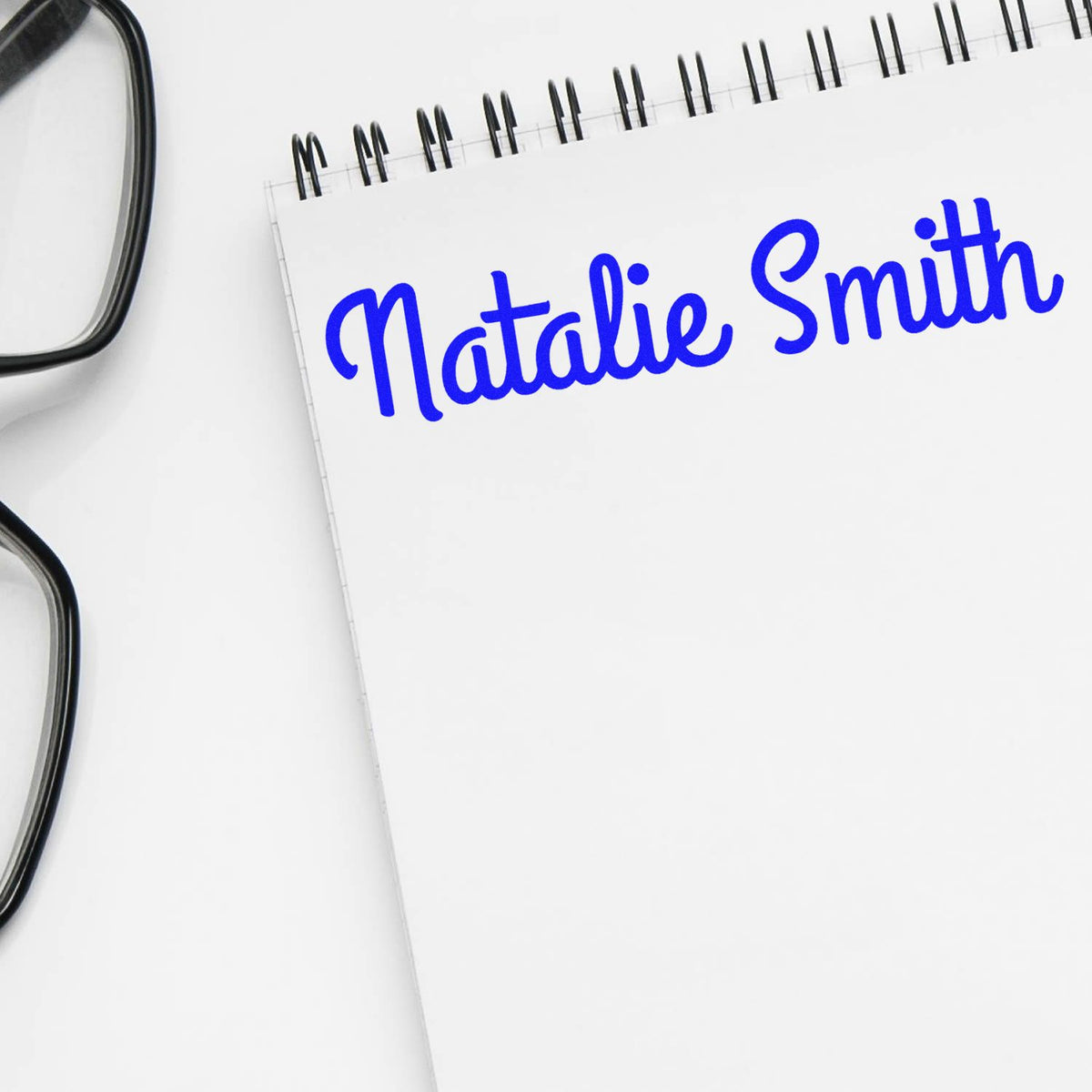 Blue custom stamp seal reading Natalie Smith on a white spiral notebook, with eyeglasses at left.
