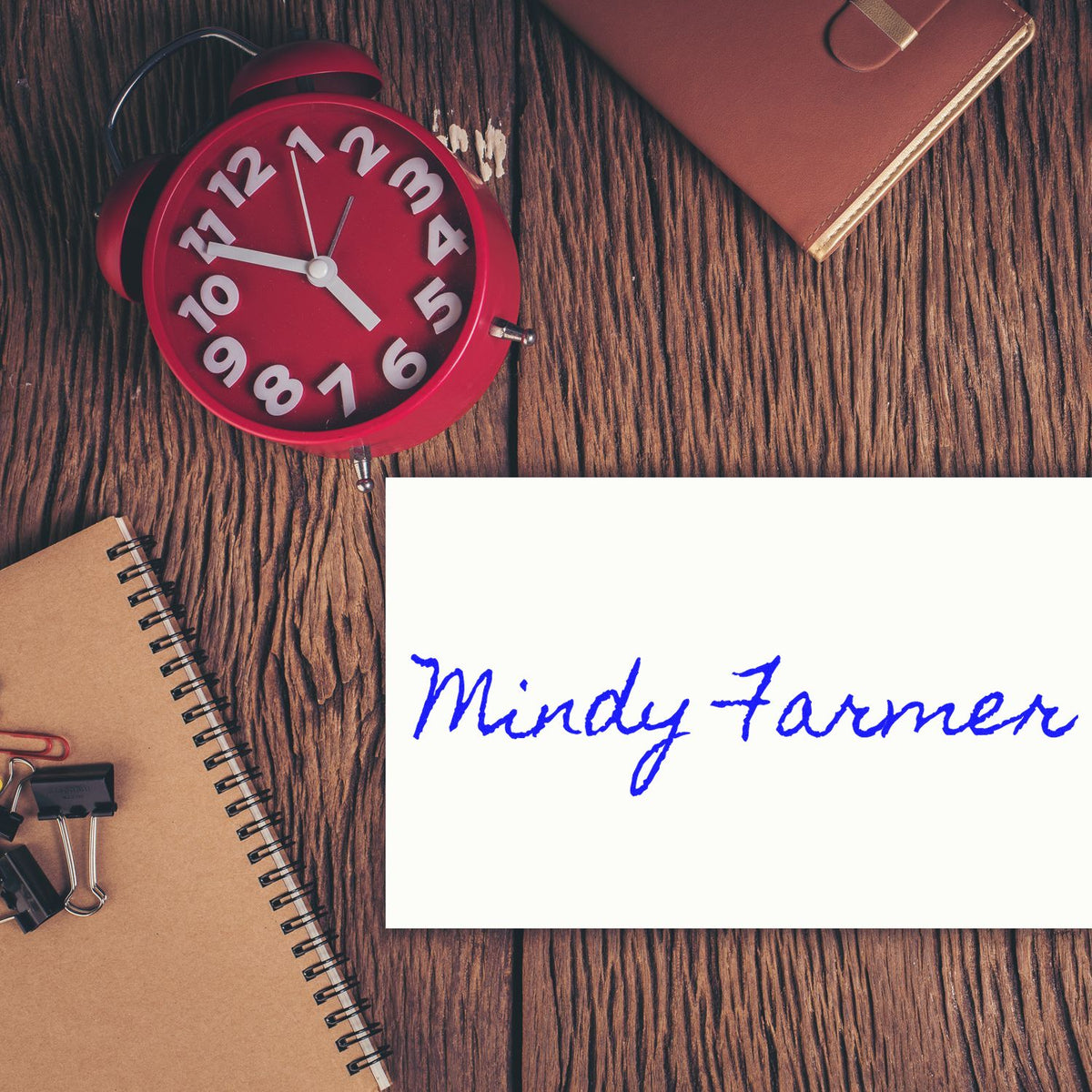 Custom stamp and seal card with blue Mindy Farmer signature, beside a red alarm clock, brown notebook, and planner on wood.