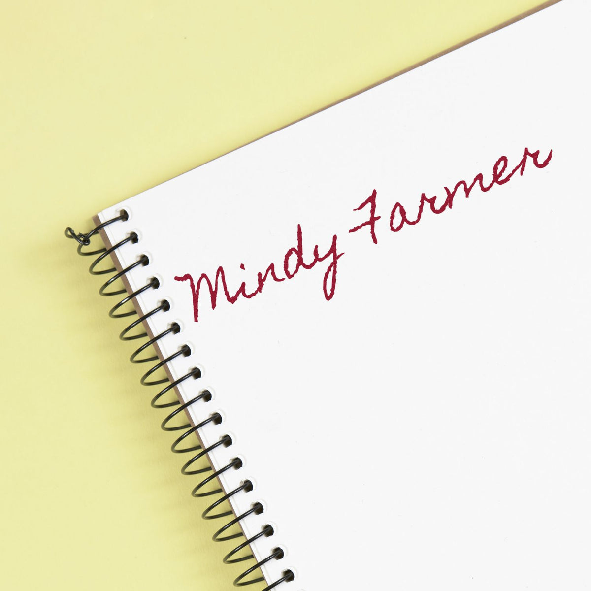 White spiral notebook on pale yellow background with red custom stamp-style script reading Mindy Farmer