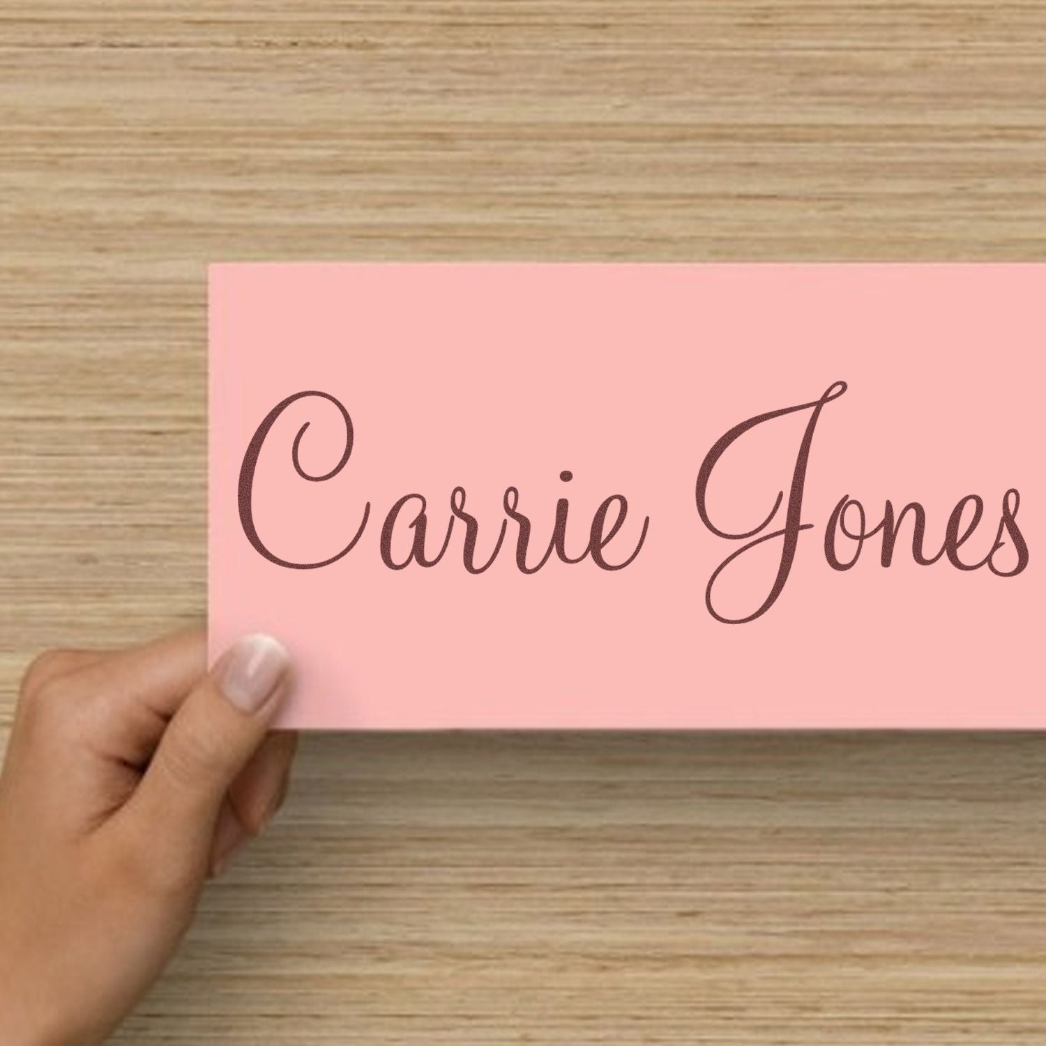 Pink custom stamp and seal sample with dark brown script reading Carrie Jones, held over a light wood surface.
