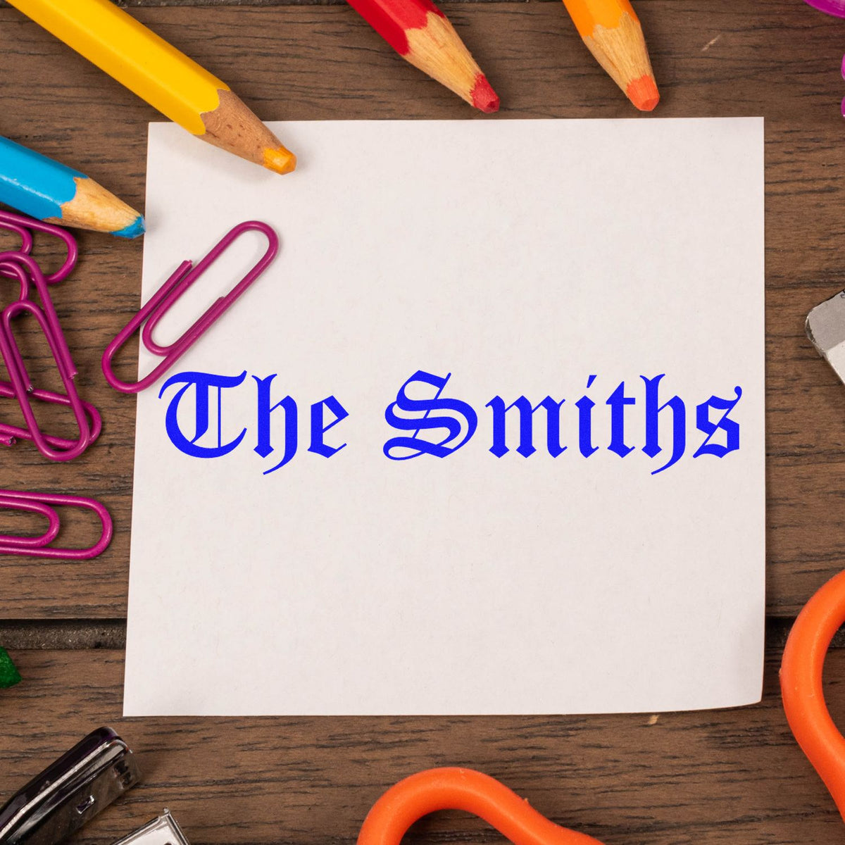 Blue custom stamp and seal reading The Smiths on white paper, framed by colorful pencils and magenta paperclips.