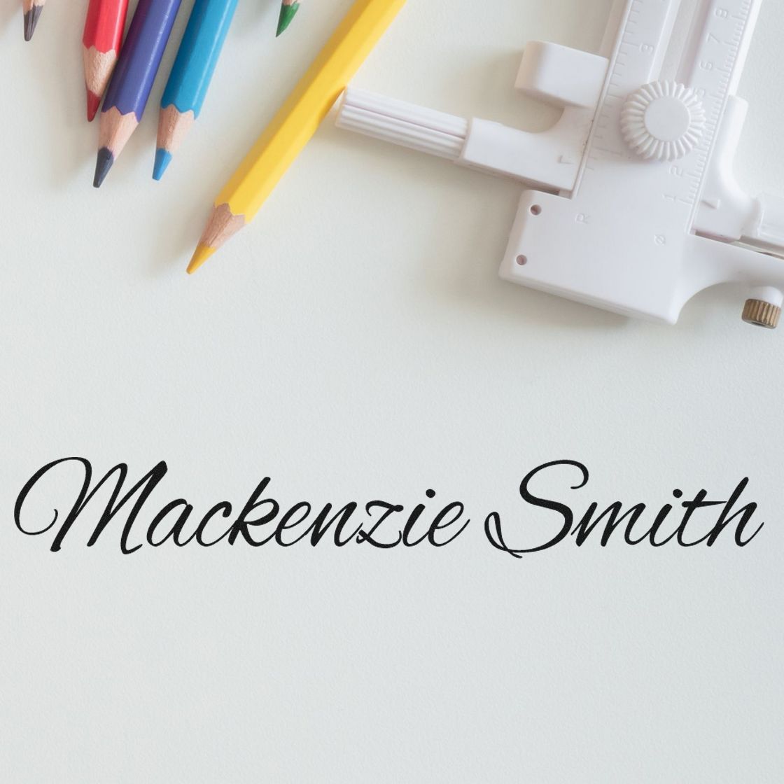 White custom stamp and seal tool with black script Mackenzie Smith, beside colorful pencils on a white background.