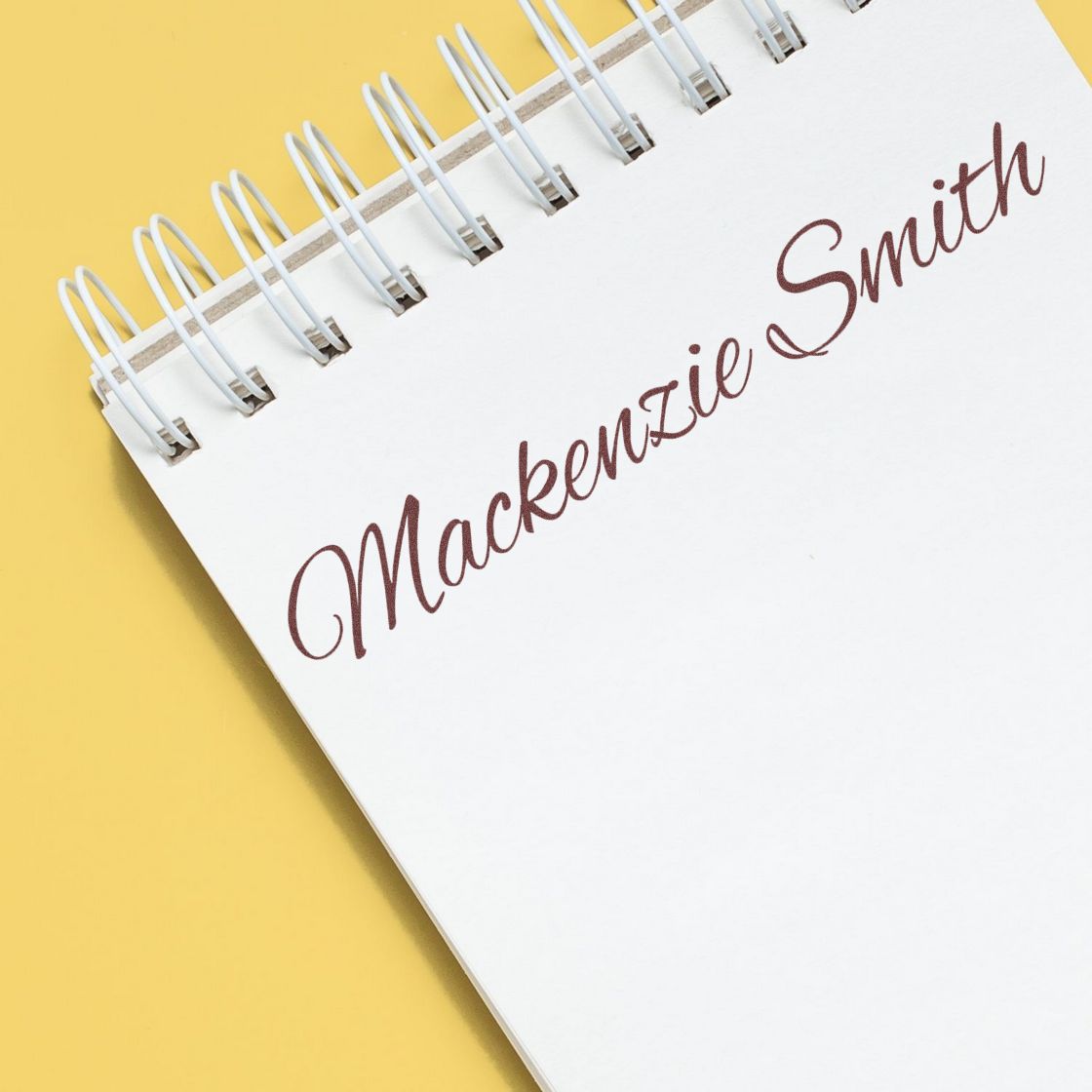 Brown "Mackenzie Smith" custom stamp/seal on a white spiral notebook over a yellow background.