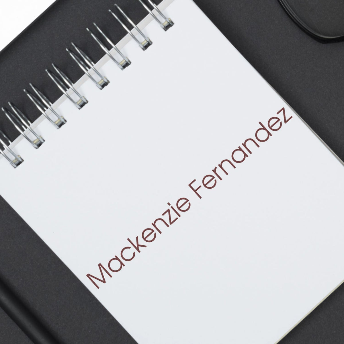 White spiral notebook with brown Mackenzie Fernandez text, shown as a custom stamp and seal product on a black background.