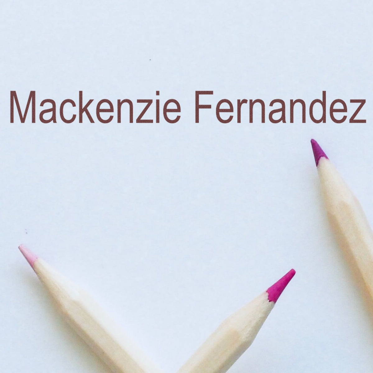 Brown Mackenzie Fernandez stamp on white paper, framed by three pink-tipped pencils.