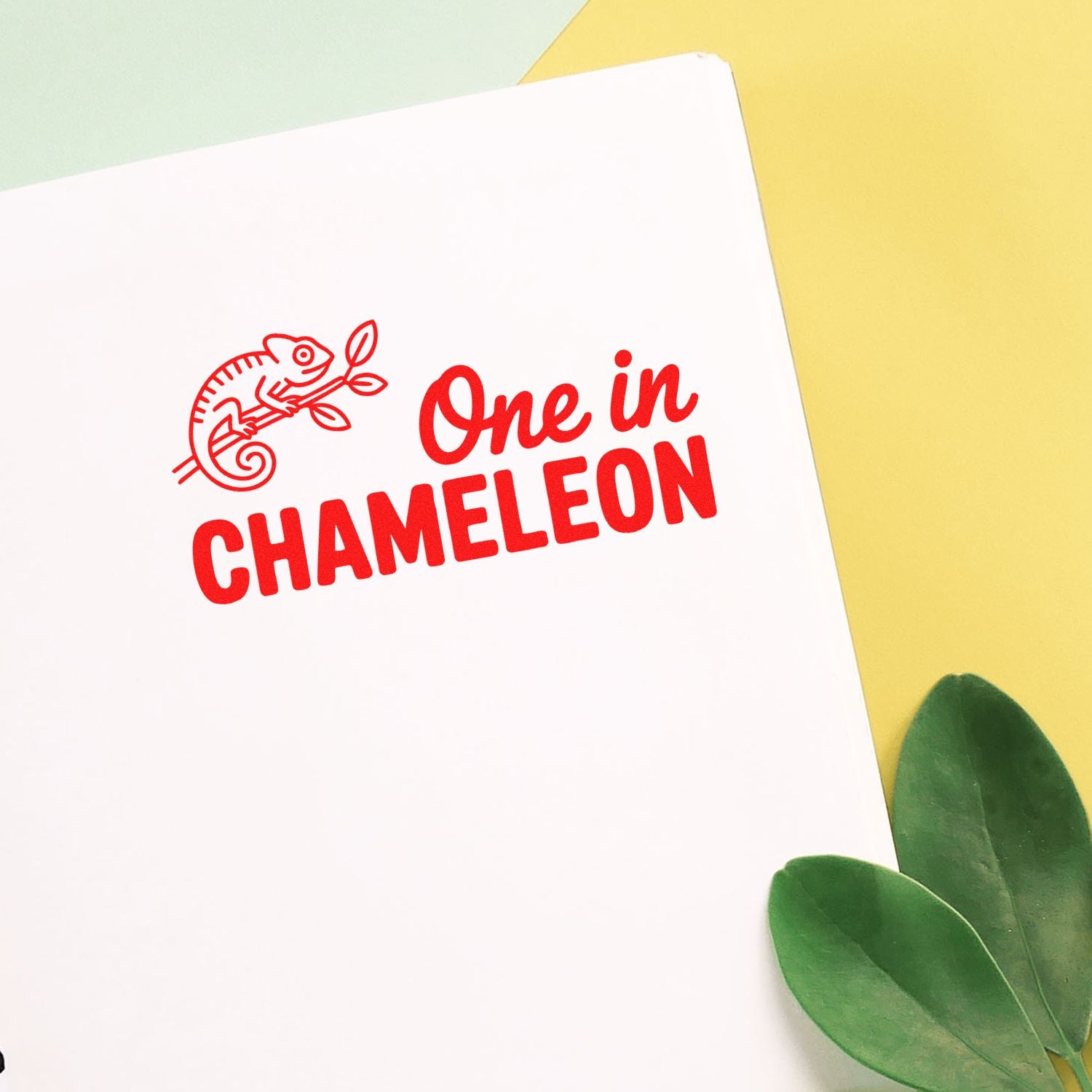 Red chameleon stamp and seal on white paper with One in CHAMELEON text, against a yellow background and green leaves.