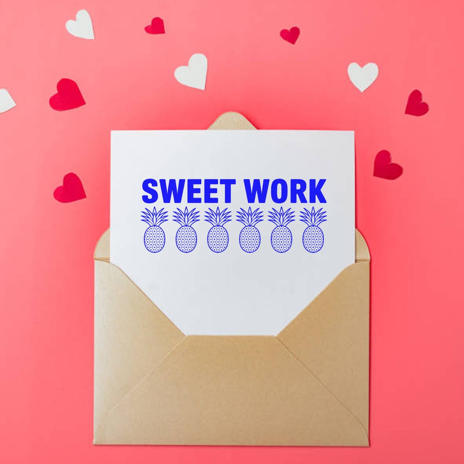 Beige envelope with white card reading SWEET WORK, blue pineapple stamp seals, on pink background with heart confetti.