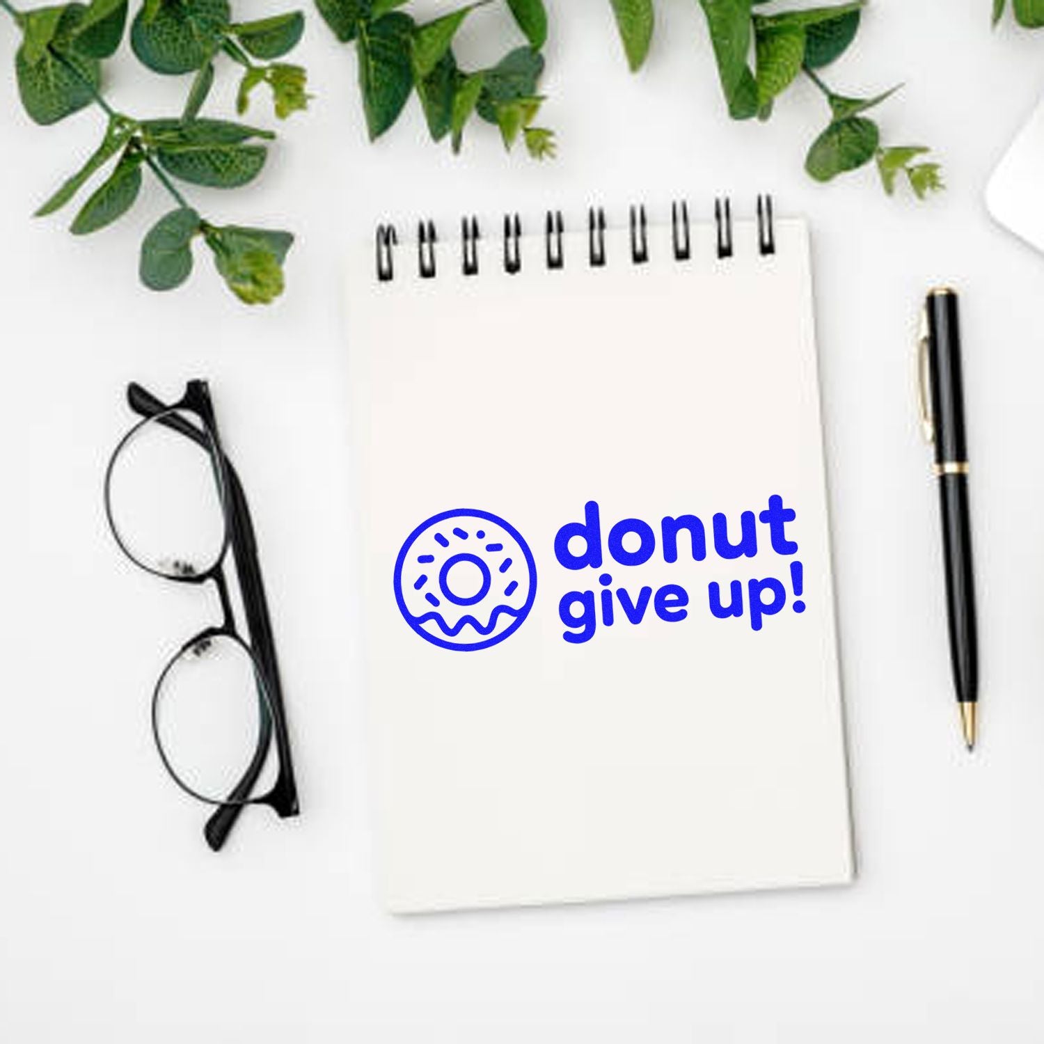 Blue donut seal stamp with donut give up! on a white spiral notebook, beside black glasses, pen, and green leaves.