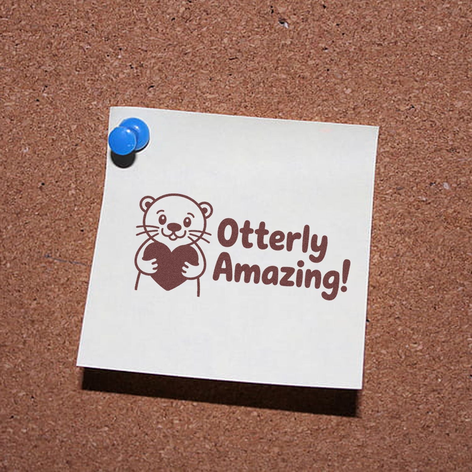 Brown otter stamp holding a heart with Otterly Amazing! on white paper pinned to a corkboard with a blue pushpin.