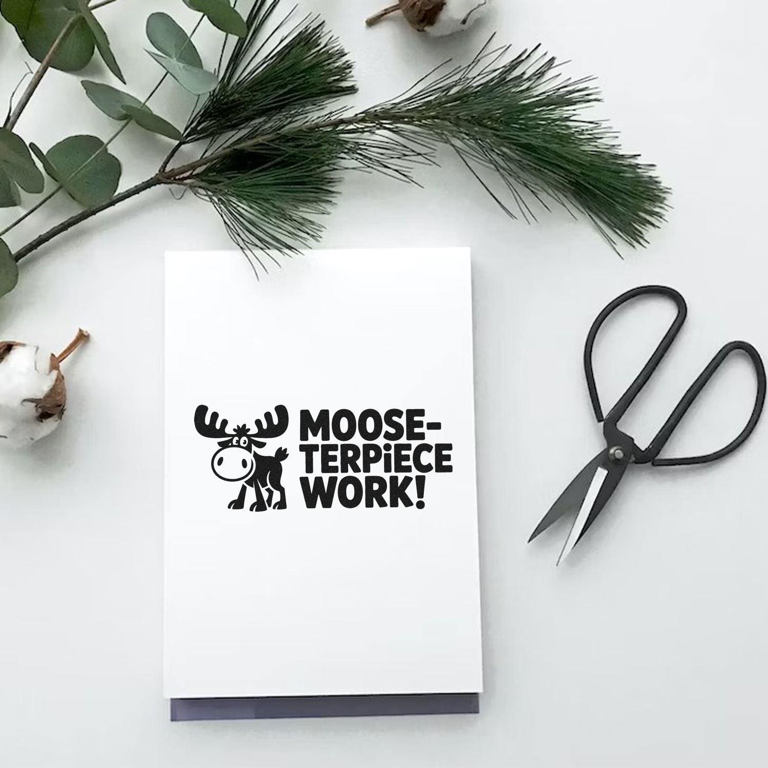 White card with black custom stamp and seal reading Moose-terpiece Work!, beside greenery and black scissors.