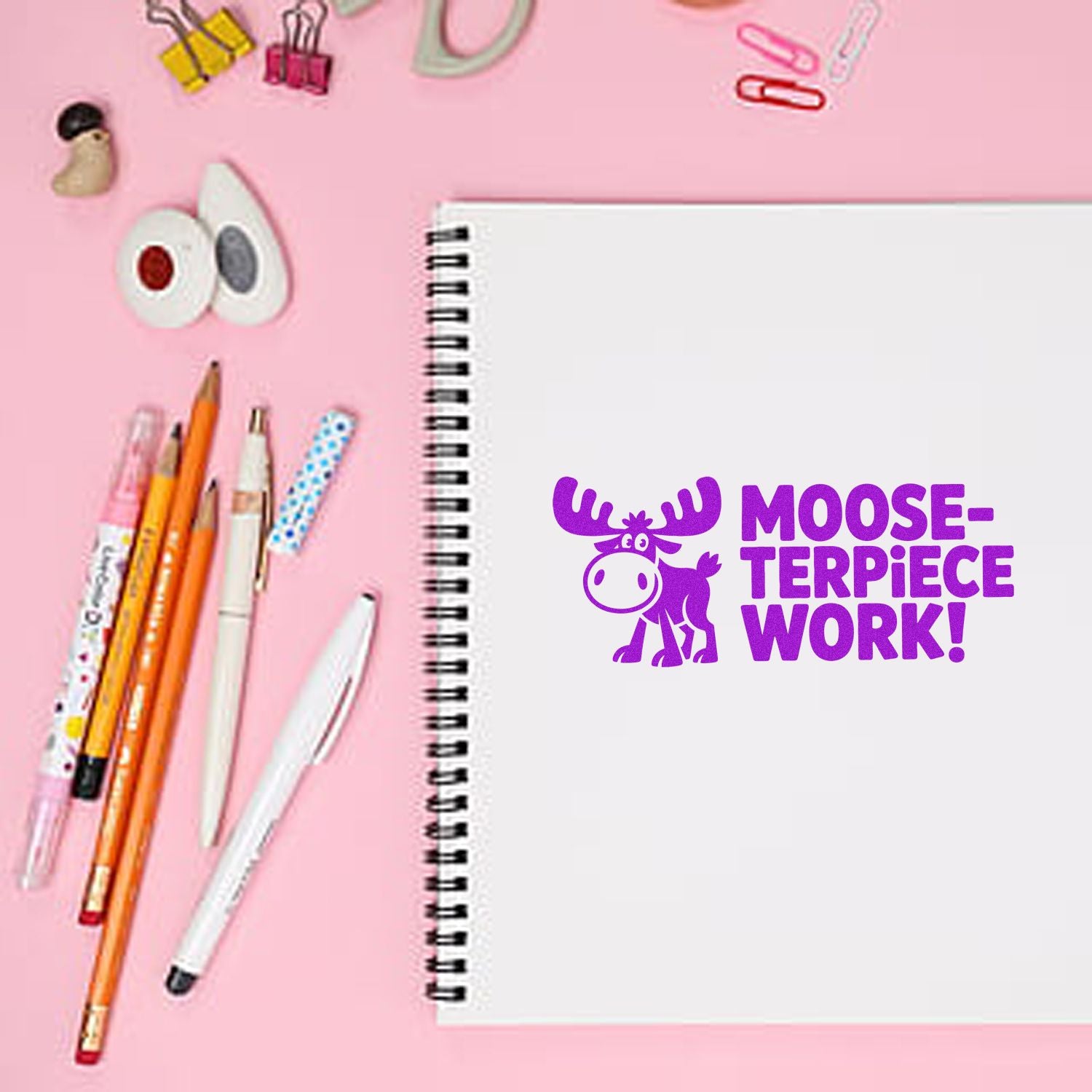 Purple moose stamp seal on a white spiral notebook, with pink background and colorful pens, clips, and stationery.