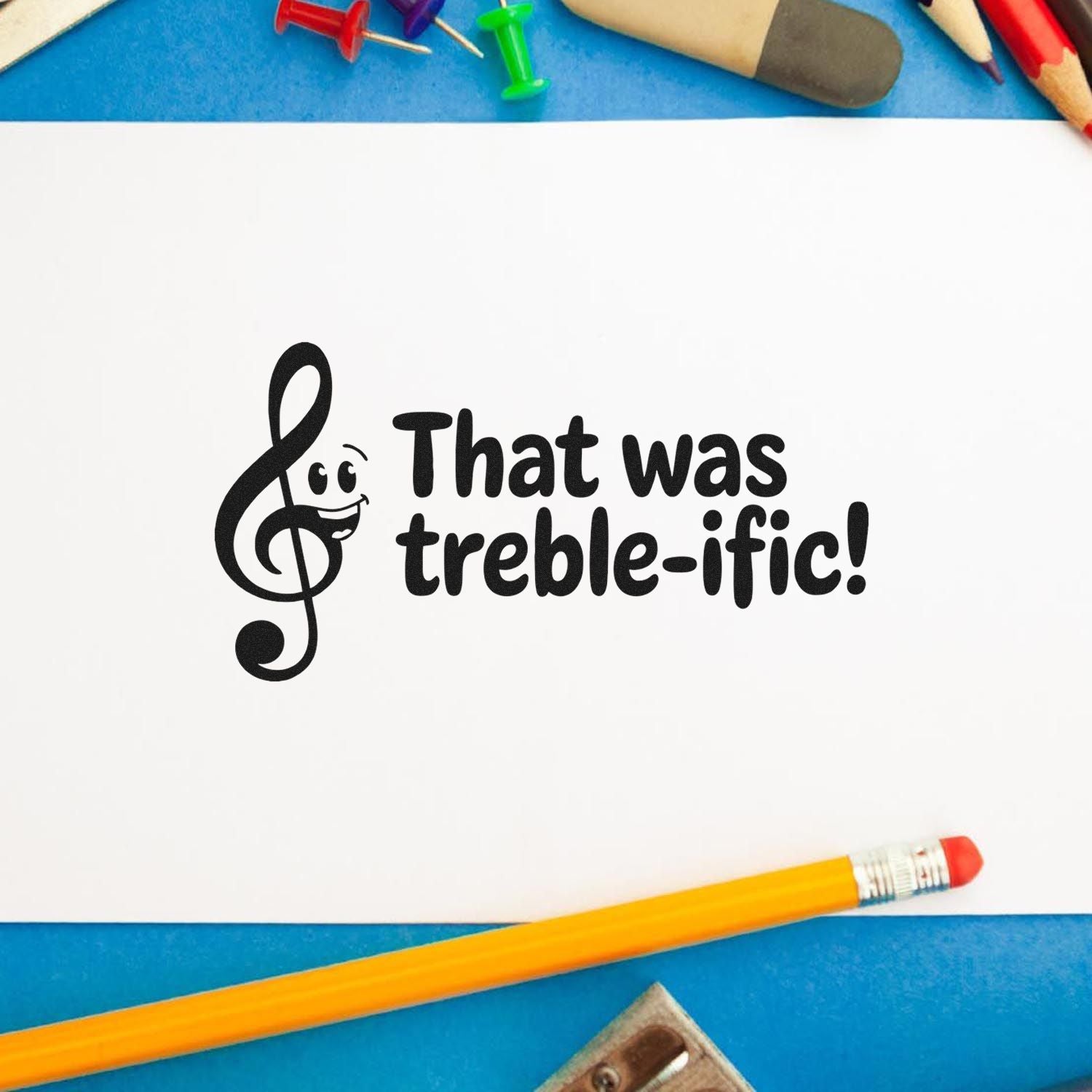Black treble-clef stamp with That was treble-ific! on white paper, surrounded by colorful pencils and push pins on blue background.