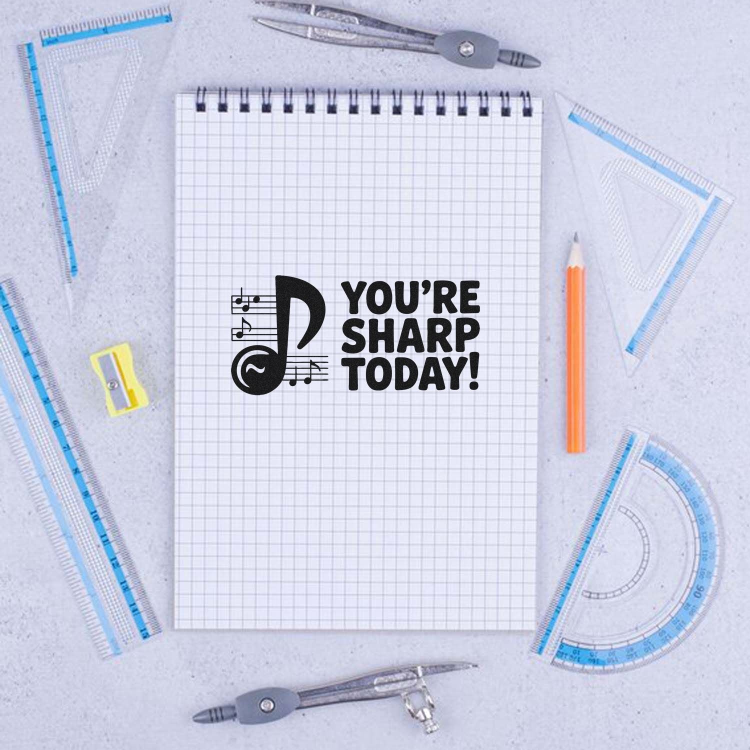 Black music note stamp with Youre Sharp Today! on graph paper notebook, surrounded by blue rulers, gray compasses, and orange pencil.