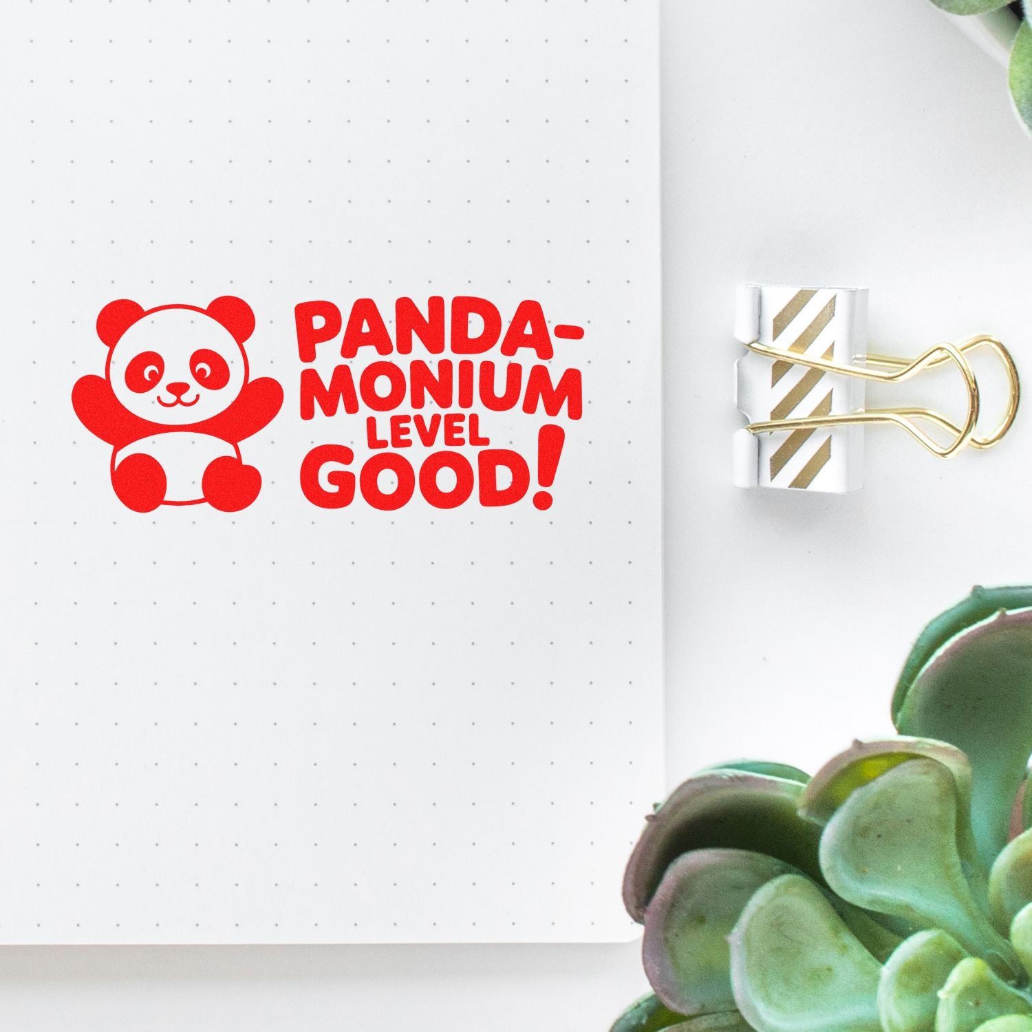 Bright red panda stamp reading PANDAMONIUM LEVEL GOOD! on dotted notebook page, beside a white-and-gold striped seal clip.