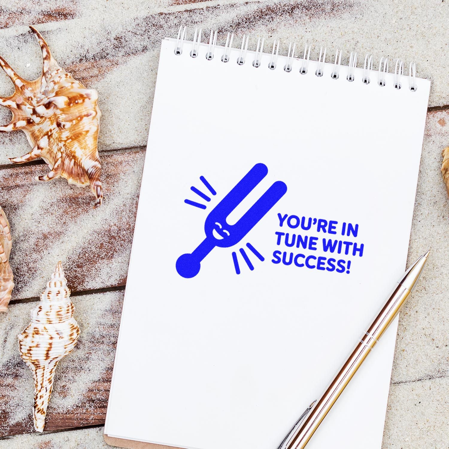 Blue stamp seal with Youre in tune with success! on white spiral notebook, beside seashells and a gold pen.