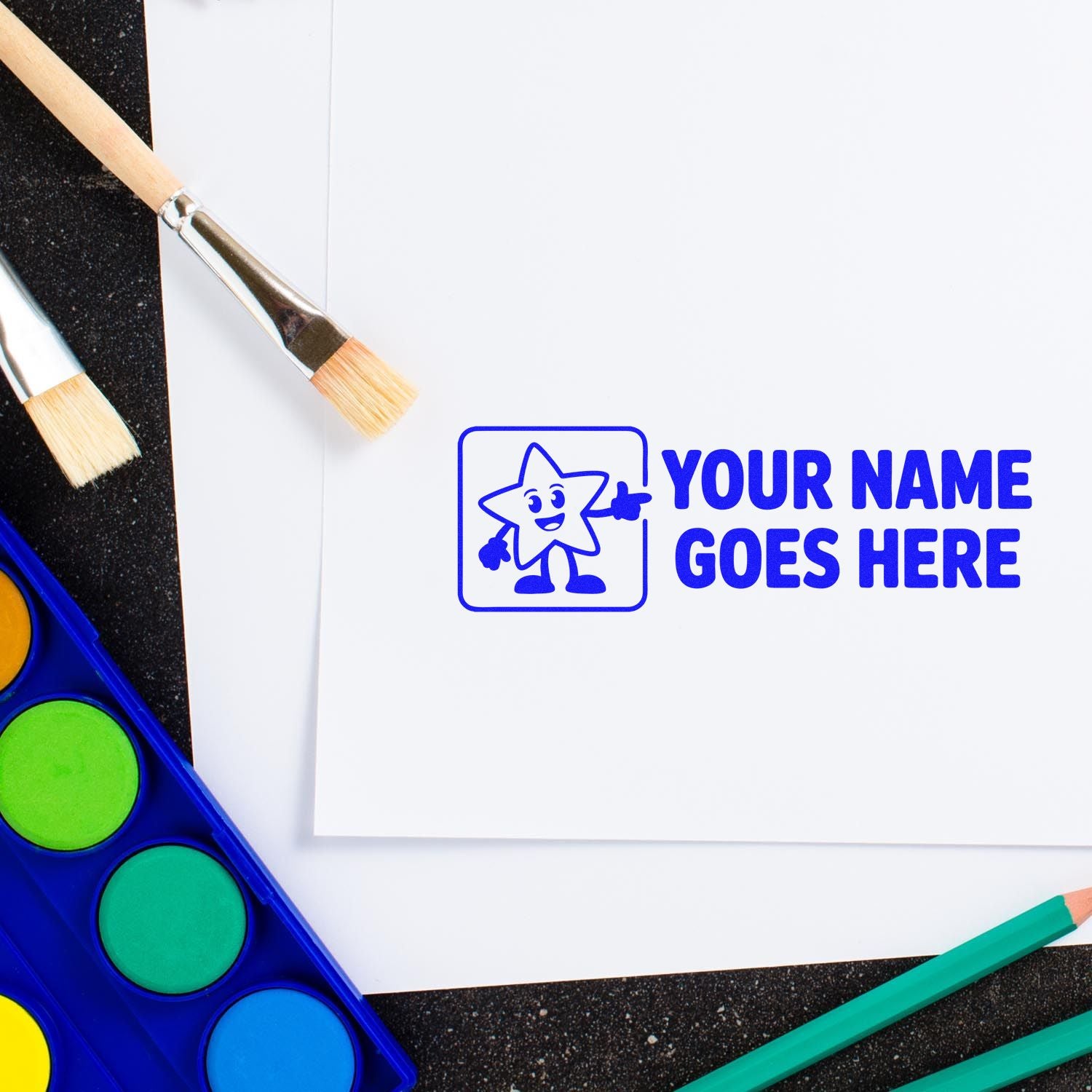 Blue custom stamp and seal with star mascot and Your Name Goes Here on white paper, beside paintbrushes and watercolor palette.